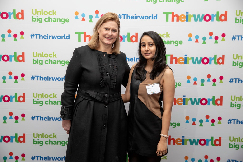 Palak Sharma stands with Sarah Brown smiling in front of a wall featuring the Theirworld logo repeated